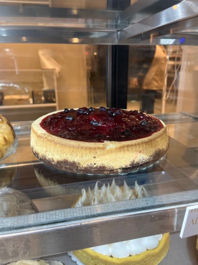 A close-up shot depicts a berry cheesecake on display inside a glass-fronted refrigerated display case. Other pastries are visible behind and below the cheesecake, including a lemon meringue tart. The location is likely a bakery or cafe in an urban setting, possibly San Fernando del Valle de Catamarca, Argentina.