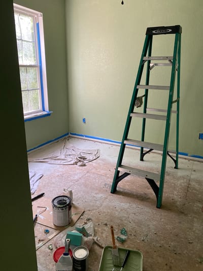 The image depicts an interior scene that appears to be undergoing renovation or painting. A green Werner ladder stands prominently in the center of the room, leaning against a light green wall. The floor is covered with a drop cloth and has scattered paint cans, rollers, brushes, and other painting supplies. Blue painter's tape is visible along the baseboards and around a window. The room has a window with multiple panes, through which trees and foliage are visible, suggesting it is daytime. An electrical outlet is visible on one of the walls. The overall impression is that of a room in the process of being painted, with materials laid out and protection in place. The location is identified as Elizabeth City, United States.