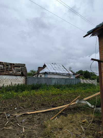 The image, taken in Pysarivka, Ukraine, depicts a damaged outdoor residential area under an overcast sky. In the foreground, the ground is covered with dark soil, sparse green vegetation, and scattered debris including broken wooden planks, a section of bent corrugated metal, and various wires. A grey, corrugated metal fence stretches across the midground, separating the foreground from two houses. The house on the left exhibits a severely damaged roof with exposed wooden rafters and fragmented or absent grey asbestos-cement sheets. Its wall is light-colored brick or painted masonry. To the right, another house features a gabled roof partially covered with silver corrugated metal sheets, and a wooden ladder is propped against it. A satellite dish is mounted on this roof. The house's walls are light blue and white. Multiple dark power lines are visible traversing the grey, cloudy sky in the upper portion of the frame. No individuals are present.