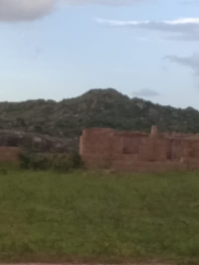 The image depicts an outdoor scene under a cloudy sky. In the foreground, a field of green grass extends towards a partially constructed building made of reddish-brown bricks. The building appears to be an unfinished structure with visible walls and openings. Behind the building, a densely vegetated hillside rises, forming a natural backdrop. The overall impression is a rural or semi-rural environment, possibly in the city of Barakin Acha, Nigeria, given the context. The image quality is somewhat blurry, making it difficult to discern finer details. There are no visible people or specific actions occurring in the scene.