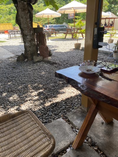This image depicts an outdoor seating area at a coffee shop in Aluepiet, Indonesia. In the foreground, a woven chair is partially visible to the left, and a rustic wooden table with a live edge occupies the right side of the frame. On the table, an empty glass with remnants of a drink sits on a saucer, alongside a smartphone and charging cables plugged into an electrical outlet mounted on a yellow pillar. The ground is covered with small, gray pebbles, with a concrete path visible in the lower foreground. Behind the table, a large tree stands near a decorative stone arrangement. In the background, picnic tables, umbrellas, and a dark SUV are visible. Further in the distance, a building with people sitting inside and lush greenery suggest the broader environment of the establishment. The lighting indicates it is daytime.