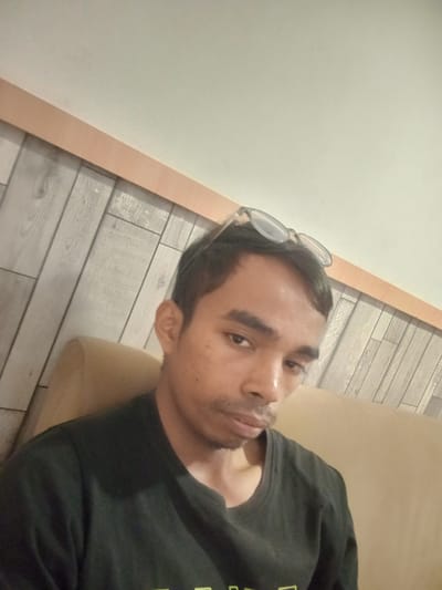 This is a selfie of a person sitting on a couch. The person is wearing a black t-shirt and has glasses perched on top of their head. They have dark hair and a light complexion. The background features a wall with a wood-paneled pattern and a light-colored ceiling. The lighting suggests it could be daytime or evening indoors. The person's expression is neutral. There is no visible text in the image. The location is identified as Larangan, Indonesia.