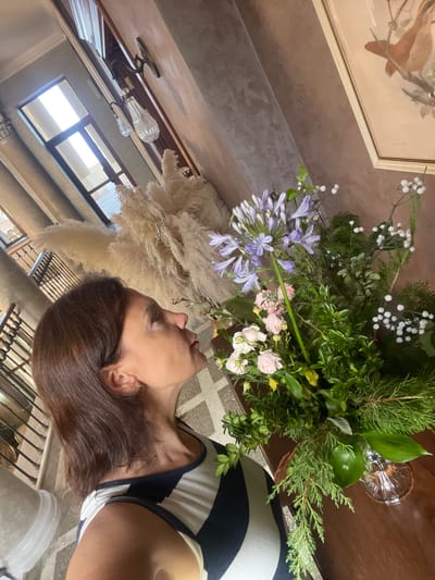 A woman is seen from the chest up, looking towards a floral arrangement on her right. She has shoulder-length brown hair and is wearing a black and white striped top. Her head is tilted upwards, and she appears to be smelling the flowers. The floral arrangement includes purple agapanthus, pink and white roses, and various green foliage, some of which are wispy and delicate, others are more structured. A vase is visible at the bottom right, holding the flowers. The background shows a textured wall, possibly painted or plastered, in muted tones of pink and beige. To the left of the woman, there is a large, fluffy decorative element, possibly dried pampas grass. Further in the background, a staircase with a black metal railing is visible, along with architectural details that suggest an interior space with large windows and possibly an atrium or a grand hallway. The overall lighting suggests daylight. The image does not provide enough information to determine the specific location within the city of Gagra, Abkhazia, other than it is an indoor scene.