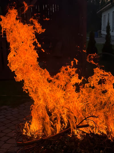 The media file shows a close-up, dramatic view of a fire. The flames are a vibrant orange, with hints of yellow and red, creating a dynamic and swirling pattern against a dark background. The fire appears to be consuming some dry vegetation, possibly branches or brush, which are visible on the right side of the frame, partially obscured by the flames. There is a hint of a chain-link fence in the lower right corner, suggesting the fire might be near a property boundary.
The overall scene is dark, with the light emanating solely from the fire. This suggests the image was taken at night or in very low light conditions. The texture of the flames is detailed, showing the movement and intensity of the fire. Scattered throughout the darker areas are small, bright specks, which could be embers or sparks rising from the blaze. The background is indistinct, but a blurry, dark shape with horizontal lines in the upper left corner might be a building or structure, possibly reflected in a surface.
There is no visible text or clear indication of people. The emotion conveyed is one of intensity and perhaps a touch of danger due to the destructive nature of fire. The context of Dreiliņi, Latvia, provides a geographical location for the event, but no specific local cues are apparent within the image itself beyond the general impression of an outdoor setting.