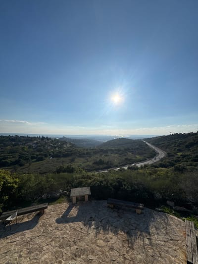 The image depicts a panoramic view from a high vantage point within the Menashe Regional Council, Israel, under a clear, bright blue sky with the sun visible in the upper right quadrant, radiating strong sunbeams. The foreground consists of a stone-paved observation area. Two rustic wooden benches are positioned on this platform, one on the left and one on the right, facing generally towards the vista. A small, white plastic chair is situated next to the left bench. Centrally located on the platform is a low, rectangular stone or concrete plaque/table. Long shadows are cast across the platform, indicating the time of day and the sun's position.
Beyond the platform, a verdant landscape of rolling hills covered in dense green vegetation, primarily shrubs and trees, extends into the distance. A winding asphalt road traverses the midground, curving from the right side of the frame towards the left, with several vehicles visible as small specks, indicating traffic flow. To the left of the road, nestled amidst the greenery, is a cluster of buildings comprising a small settlement or village. In the far background, a body of water is visible on the horizon, appearing as a reflective strip under a hazy sky, possibly the Mediterranean Sea. No individuals are visible on the observation platform or within the immediate foreground.