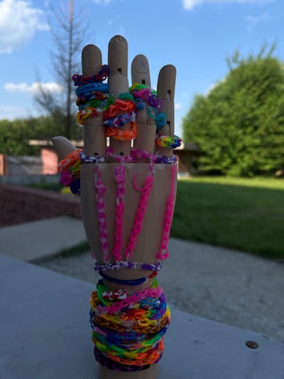 This is a close-up, rotated photograph of a wooden mannequin hand adorned with numerous colorful loom band bracelets. The hand is positioned on a flat, light-colored surface. The bracelets are made from small, elastic rubber bands and are woven into various patterns, creating a vibrant and textured display. Some bracelets are simple bands, while others are more intricate, featuring charms or layered designs. The loom bands come in a wide spectrum of colors, including pink, blue, green, orange, yellow, purple, and rainbow patterns.

The background suggests an outdoor setting. The upper left portion of the image shows a bright blue sky with scattered white clouds, indicating fair weather. A large, out-of-focus green tree is visible behind the hand, and to the right, a blurred green area likely represents more foliage or grass. The lighting suggests it is daytime, likely late morning or early afternoon, with sunlight casting subtle shadows. There is no visible text in the image. The overall impression is one of playful creativity and a celebration of color, likely captured in a backyard or park setting in Dzerzhinsk, Russia, given the provided location context.