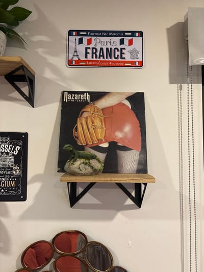 The image shows a shelf with various items. The shelf is made of wood and has black metal brackets. The items on the shelf include a record album, a Paris France license plate, and a small lizard figurine. The record album is titled "Nazareth - The Catch." The license plate is red, white, and blue and has the words "Fluctuat Nec Mergitur" and "Paris France" written on it. The lizard figurine is green and has a long tail. The items are positioned on the shelf in a symmetrical way. The location is not clear but the coordinates provided (latitude: 20.711, longitude: -100.442) indicate a location in the city of Juriquilla, Mexico. The record album features a woman wearing red leather shorts and fishnet stockings, holding a baseball glove. The woman’s face is not visible. There is a plant on the left side of the image, just above the shelf, but it is not visible in its entirety.