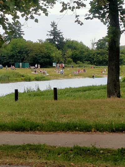 The image depicts a sunny day at a riverbank in Nosivka, Ukraine. A group of people are scattered along the grassy bank, enjoying leisure activities. Several individuals are sunbathing on towels and blankets, while others are wading or standing in the shallow water. Some people are gathered in small groups, conversing and relaxing.

In the background, there are green changing rooms or small structures, and beyond that, a dense line of trees forms the backdrop. A dark-barked tree trunk dominates the right side of the frame, its branches partially obscuring the sky. The sky itself is overcast, suggesting a hazy or cloudy day, despite the apparent brightness. The river is calm and reflects the subdued light.

Two dark posts are visible in the foreground, planted in the grass near the river's edge. A paved path runs through the lower portion of the image, with grass on either side. The overall atmosphere appears peaceful and relaxed, with people enjoying a day outdoors by the water. There is no visible text in the image.