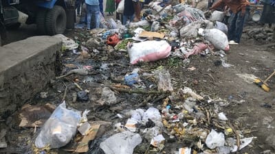This image captures a highly cluttered outdoor scene in Rubavu, Rwanda, characterized by a substantial accumulation of diverse waste materials. The foreground and midground are dominated by dense piles of mixed refuse, including decaying organic matter, such as fruit and vegetable scraps, intermingled with numerous plastic bags, torn cardboard, and general debris.

In the lower-left, a dark, murky liquid has pooled next to a weathered concrete curb. To the immediate left, the lower section and large tires of a blue, heavy-duty truck are partially visible.

Multiple individuals are present in the background. One person, wearing a brown-orange top and blue jeans, is observed bending over a large waste pile in the mid-right portion of the frame. Other individuals are partially discernible further back, near the truck and additional waste accumulations. A yellow and black-bristled broom lies on the uneven, soiled ground in the far-right foreground, suggesting recent or ongoing cleaning activity. The overall environment appears unsanitary and unmanaged, with the ground visibly dirty and uneven due to the extensive refuse.