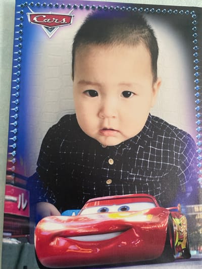 The image is a close-up photograph of a young child in a checkered shirt, likely posed in a studio setting. The child's face is the central focus, with a slightly blurred background. The photograph has a decorative border on the left and right sides, featuring a pattern of blue diamond shapes. The upper portion of the image includes a "Cars" movie logo. In the foreground, a red toy car resembling Lightning McQueen from the "Cars" movie is prominently displayed, partially obscuring the lower part of the child. The overall impression is a themed portrait. There is no indication of the location being the city of Бескөл ауылдық округі, Kazakhstan.