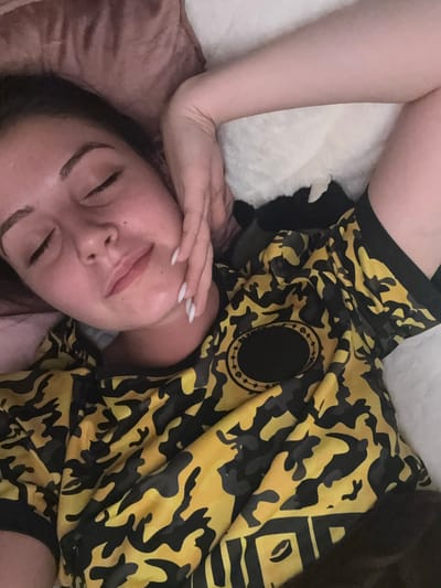 A young woman with closed eyes and her hand on her cheek is lying down, possibly in bed. She is wearing a yellow and black camouflage patterned shirt with a circular emblem on the chest. The background includes a soft, reddish pillow and a white, fluffy surface. The scene appears to be an indoor setting.