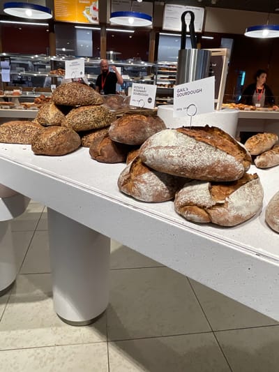 This image depicts the interior of Gail's Bakery, located at 2 The Broadway, Crawley, RH10 1DS, United Kingdom.
In the foreground, a prominent white display counter showcases a substantial collection of freshly baked artisan bread loaves. The bread is arranged in multiple stacks and piles across the counter's surface. Distinct varieties are identified by white signs on metal spikes: "SEEDED SOURDOUGH," "POTATO & ROSEMARY SOURDOUGH," and "GAIL'S SOURDOUGH" (priced at 650g £4.90). The loaves exhibit varied crusts, some coated in white flour, others richly browned and deeply scored, and several are visibly topped with seeds.
In the midground, behind the bread display, two staff members are visible. On the left, a bald male with a beard, wearing a dark shirt and a lanyard, stands behind a counter, gesturing with his right hand near his ear. Further to the right, a female staff member in a dark top and light-colored apron stands behind another counter, looking towards the right side of the frame. Various items are on display behind glass counters, including pastries and smaller bread items. A tall, stainless steel utensil holder with dark tongs is positioned prominently.
The background features dark reddish-brown walls and sections of glass partitioning, revealing a commercial kitchen or preparation area with stainless steel surfaces and shelving. Overhead, several blue-rimmed white pendant lights illuminate the space. Menu boards are visible, reading "FROM THE KITCHEN," "FROM THE FRIDGE," and "FROM THE BARISTA," along with a yellow sign advertising "SUMMER" items.
The floor is light-colored tile with visible grout lines and some minor dark speckling. The white display counter is supported by thick, white cylindrical columns.