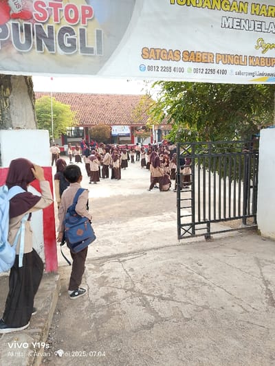 The image shows a school gate in Kecamatan Haurgeulis, Indonesia, located at latitude -6.42 and longitude 107.971. A large group of students in brown uniforms are dispersing from the school grounds. Two students, one with a blue backpack, stand by the gate, with a third student standing behind them with their arm raised. The gate is made of black metal and opens inward. The school building is visible in the background. Above the gate is a sign that reads "STOP PUNGLI SATGAS SABER PUNGLI KABUP" with phone numbers listed underneath. The sign is attached to a larger sign, but only a portion of it is visible, displaying the words "TUNJANGAN HARI MENJELA". The school grounds are paved with a grey concrete surface. The trees surrounding the school are partially visible in the background.