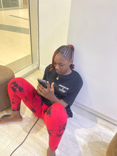 A young woman with braided hair is sitting on the floor indoors, looking at her phone. She is wearing a black t-shirt with white text that says "WARDROBE & CLOSET" and red leggings with a black abstract pattern. Her legs are spread apart, and her feet are bare. To her left is a brown ottoman, and behind her is a white wall. To her left, through a glass partition, a tiled floor is visible in what appears to be a public or commercial space, with tiled flooring and soft lighting. The overall environment suggests an indoor setting in Unguwar Sarki, Nigeria.