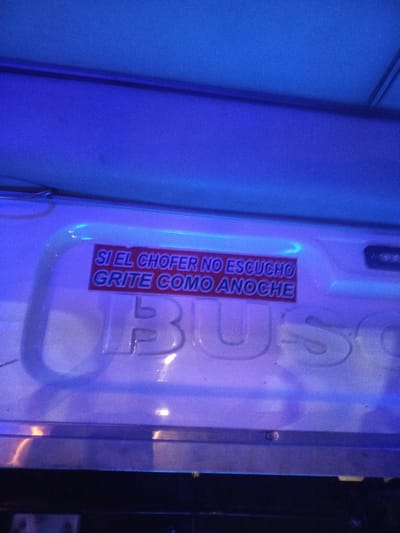 The image shows the interior of a bus. A sign reads "Si el chofer no escucho grite como anoche", which translates to "If the driver doesn't hear, scream like last night". The word "BUS" is displayed prominently on the vehicle. The location is unknown.