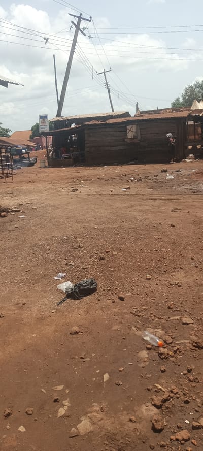 The image depicts an outdoor scene in Oturkpo, Nigeria. The foreground shows a dirt ground littered with trash, including a black plastic bag and a plastic bottle. In the midground, there's a wooden structure with a corrugated metal roof. People are visible inside the structure. Power lines are strung overhead, supported by utility poles. In the background, there's a sign for a hotel and additional structures.