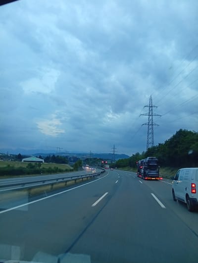 The image depicts a highway scene in Ittigen, Switzerland, under a cloudy sky. The perspective is from a vehicle moving along the highway. In the foreground, the asphalt road is visible with white lane markings. To the right, a white van is driving in the same direction. Further ahead on the right lane, a car carrier truck is transporting multiple cars. To the left of the highway, there's a grassy embankment with a metal guardrail. In the middle distance, on the left side of the highway, a building with the text "TIVOLI" is visible, along with construction cranes and trees. In the background, rolling hills and mountains are present. Tall electricity pylons with power lines are prominent in the scene, one of which is particularly large and central. The overall lighting suggests it's either early morning or late evening.