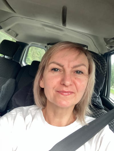 A woman with blonde hair and blue eyes smiles at the camera while sitting in the passenger seat of a vehicle. She is wearing a white t-shirt and a seatbelt is visible across her chest. The interior of the car, including headrests and a portion of the ceiling, is visible. Outside the car window, green trees and foliage can be seen, suggesting a rural or suburban setting. The image appears to have been taken in the vicinity of Драчкаўскі сельскі Савет, Belarus, based on the provided context.