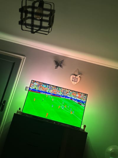 A television displaying a soccer match is the primary focus of this indoor scene. The television screen shows players in orange and blue uniforms on a green field, with advertisements for "Qatar Airways" visible. The room appears to be a bedroom or living area. A dark dresser is positioned beneath the television. Decorative items are present on the wall, including a black butterfly-shaped ornament and a wooden heart-shaped ornament inscribed with "with love". A ceiling light fixture with a cage-like design is visible above. The lighting in the room is dim and appears to be influenced by green LED strip lights, creating a moody ambiance. There is a door on the left side of the image. The scene is not located in Marten, Bulgaria.
