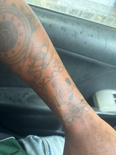 The image shows a close-up of a dark-skinned person's forearm with several tattoos. One tattoo appears to be a clock face with Roman numerals. The arm is positioned next to a dark, textured surface, possibly the interior of a vehicle. Part of a blue denim garment and green fabric are visible in the lower right corner. The scene appears to be indoors, possibly inside a car, given the interior elements and the quality of the lighting. The location in Iddo Tudunwada, Nigeria, is not discernible from the visual information.