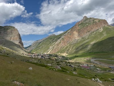 This is a landscape photograph of the village of El'tiubiu, Russia, nestled within a valley. The image features dramatic, rocky mountain slopes on either side, with a village situated in the valley floor. Lush green grass covers the foreground and parts of the mountain slopes. In the distance, a winding road with several parked vehicles, including cars and vans, is visible. Buildings with red and grey roofs are scattered throughout the village. The sky is a vibrant blue with scattered white clouds. The scene depicts a rural mountain village in a natural setting.