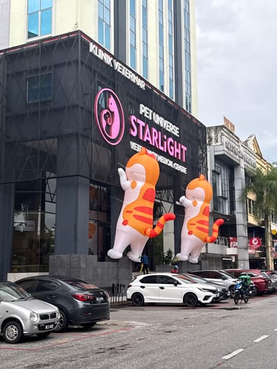 The image depicts the exterior of a veterinary clinic called "Pet Universe Starlight," located in Ampang Jaya Municipal Council, Malaysia. The building is modern with a black facade and large signage featuring the clinic's name and a pink cat logo. Two large inflatable orange and white cat figures are positioned on the roof. Parked along the street are several cars, including a white car, a dark grey car, and a silver car. A few motorcycles are parked near the white car. The street is paved, with visible road markings. Other buildings and a pharmacy are visible across the street, as well as some trees and a cloudy sky.