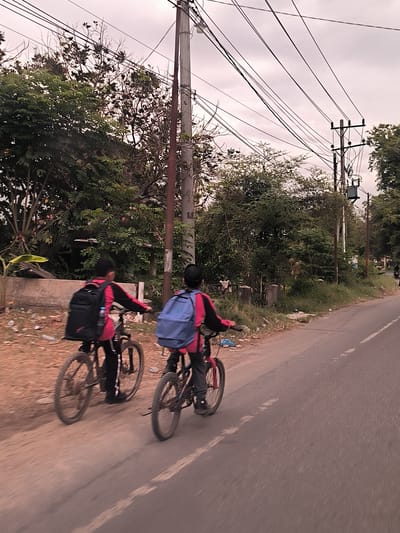 The image displays a scene of two individuals, likely children or teenagers, riding bicycles on a paved road, moving away from the camera. Both cyclists are wearing long-sleeved red or pink tops and dark trousers. The rider on the left has a black backpack, while the one on the right carries a blue backpack. They are on mountain bike style bicycles.
The road is made of asphalt, with faint white lines visible, indicating a lane or road edge. To the left of the road is a dirt shoulder with some scattered trash, including plastic bottles, and low concrete structures backed by lush green trees and foliage. To the right of the road, there are more trees, some with dense green leaves and others with bare branches, where numerous small birds are perched. Multiple utility poles, likely made of wood or concrete, line the roadside, supporting a complex network of overhead power lines. One pole is equipped with an electrical transformer.
The sky is overcast and light-colored, suggesting either an early morning, late afternoon, or a generally cloudy day. The overall lighting is soft and diffuse, giving the image a slightly desaturated, warm tint. The setting appears to be a suburban or rural area with abundant vegetation and visible utility infrastructure, aligning with the provided location context of Lhoknga, Indonesia. No visible text is present in the image.