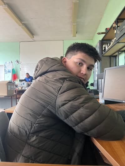 An indoor scene is depicted within a room in Cherven Bryag, Bulgaria. In the foreground, a young male with short, dark hair wears a dark olive green, horizontally quilted puffer jacket. He is seated at a light brown wooden desk, turning his upper body and head to face the camera, with his right forearm resting on the desk surface. To his right, a black computer monitor, a black keyboard, and a dark grey Dell OptiPlex computer tower are positioned on the desk.

In the midground, an adult male with short dark hair is seated at a separate light brown wooden desk to the left. He is wearing a blue long-sleeved top and is oriented towards a light-colored laptop on his desk. A small white sewing machine is also visible on his desk. Behind this individual, a Bulgarian flag on a pole leans against the wall. The room's walls are painted light green on the lower section and white on the upper section. Two whiteboards are mounted on the back wall; the one to the right of the second male displays handwritten notes. To the right of the foreground subject, a metal shelving unit contains various electronic components, including a prominent red cylindrical object on an upper shelf. Overhead, two rectangular fluorescent light fixtures with yellowed diffusers are affixed to the ceiling. A large cardboard box is visible on a table in the background, to the left of the second male. The environment suggests a classroom, workshop, or technical office setting.