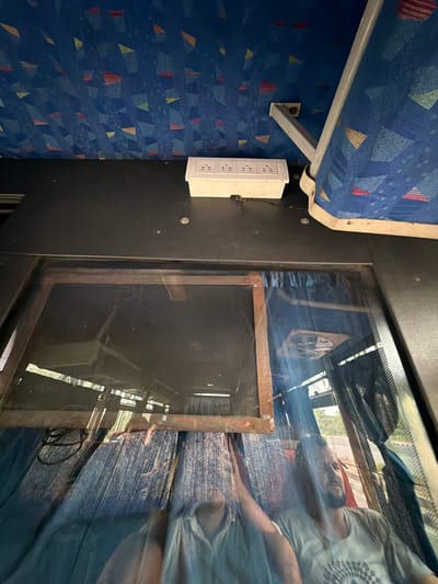 The image shows the interior of a vehicle, likely a bus, with a patterned blue fabric lining the ceiling and seats. A white power strip with multiple outlets is mounted on a dark surface. Below this, a window reflects the interior and exterior. The reflection shows two individuals, one with a beard wearing a light-colored t-shirt with a circular design, and another person partially visible. The exterior reflection suggests a road and trees, indicating the vehicle is in motion. The scene is consistent with the interior of a bus or similar passenger vehicle. The provided location, Vellanki Agros, India, is a factual detail not directly discernible from the image content itself, but it is stated as context.