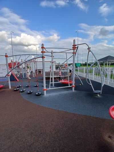This image depicts a modern outdoor children's playground with a light gray sky filled with white clouds. The playground equipment is constructed from metal with orange accents. Several children are present. One child, wearing a green shirt and light-colored pants, stands on the equipment. Another child, in a pink shirt, is on a red swing. A person in a white shirt and dark shorts stands nearby. The ground of the playground is a mix of dark gray and brown rubberized material, with a section of brown material in the foreground. In the background, there is a white fence, a covered picnic area, and a green grassy area. The scene is likely in Vorsha, Russia, given the context provided.