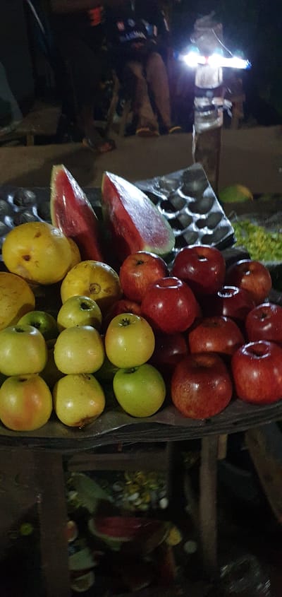 The image depicts a stall in Barakin Angundi, Nigeria, at night. The stall is laden with various fruits, including several slices of watermelon, yellow fruits that appear to be guavas or similar, and numerous red and green apples. The fruits are arranged on a dark surface, possibly a table or tray, with a textured, possibly metallic, object behind the watermelon slices. In the background, several individuals are visible, partially obscured by darkness. One person is standing and appears to be illuminated by a bright, possibly handheld, light source. The overall scene suggests a bustling nighttime market environment, with the fruits illuminated by artificial light.
