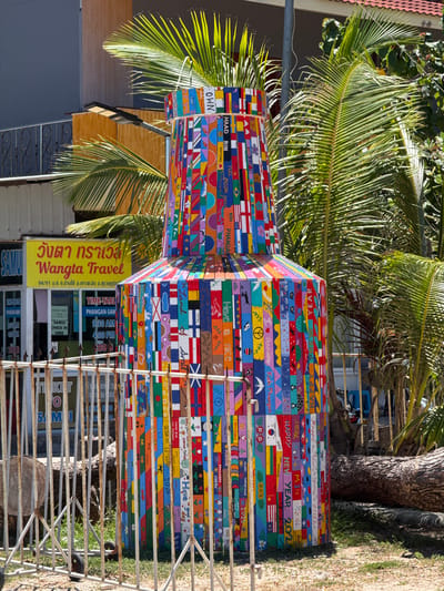 In the city of Ban Haad Rin, Thailand, a colorful, cylindrical structure, resembling a large bottle, stands prominently outdoors. This structure is adorned with numerous vertical planks, each painted with various designs. These designs include numerous national flags, such as those of Thailand, Finland, and Brazil, interspersed with text like "HAPPY NEW YEAR," "MERRY CHRISTMAS," "FULL MOON PARTY," and "KOH PHANGAN." Some planks feature decorative elements like flowers, peace signs, ladybugs, and birds. Behind the structure, a building with a yellow sign that reads "Wangta Travel" in Thai and English is visible. Palm trees with their fronds arching over the scene suggest a tropical environment. A metal fence with a rusty patina runs in the foreground, partially obscuring a large fallen tree trunk on the right side of the image. The ground is covered with gravel and grass, and sunlight casts shadows, indicating it is daytime.