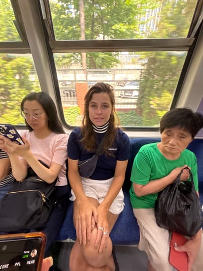 Three individuals are seated on a train in Incheon, South Korea. On the left, an East Asian woman wearing glasses and a pink shirt is looking at her phone. In the center, a white woman with brown hair, wearing a navy blue Adidas shirt and white shorts, is looking forward. On the right, an East Asian woman with black hair, wearing a green shirt and beige pants, is holding a black plastic bag and appears to be looking down. The train car has blue seats and large windows showing greenery and buildings outside. The lighting suggests it is daytime.