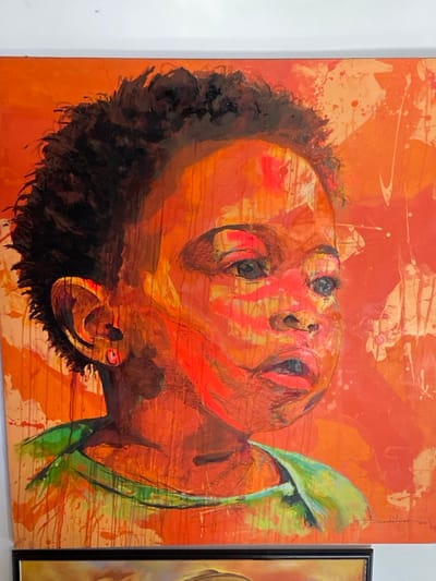 The image displays a large, vertically oriented expressionistic painting, located in the city of Ikate, Nigeria. The artwork portrays the head and upper torso of a child with dark, curly hair, gazing upwards and to the right with a slightly open mouth. The child's left ear features a small, red circular earring. The predominant color palette consists of vivid orange and red hues, characterized by prominent brushstrokes, drips, and splatters that give the painting a textured appearance. A green garment is visible on the child's upper body. The painting is affixed to a plain white wall. A portion of a second framed artwork, encased in a dark frame, is visible along the bottom edge of the image.