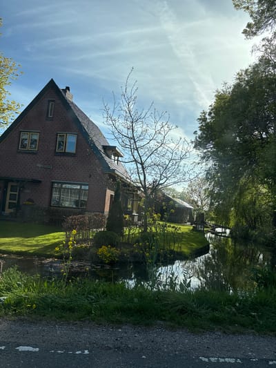 This image, when correctly oriented, presents a serene, sunlit landscape in De Lier, Netherlands. The central subject is a two-story brick house with a dark gabled roof, featuring multiple light-framed windows and visible skylights or dormers. A prominent, large front window displays a distinctive white grid pattern, and faint, illegible white text is inscribed on the brick wall below it.
Adjacent to the house, a calm canal or narrow river runs, perfectly reflecting the bright blue sky and the surrounding verdant trees. The banks of this waterway are lush with dense green grass, various plants, and clusters of vibrant yellow flowers. In the distance across the water, a small structure, possibly a bridge or another building, can be seen.
The scene is framed by abundant vegetation. Tall, leafy trees line the canal, while a deciduous tree with mostly bare branches stands near the house, suggesting early spring. Well-maintained green lawns spread around the residence. A narrow, dark asphalt road, with faint white markings, occupies the right edge of the frame. The sky is bright blue with scattered, wispy white clouds, indicating a clear, sunny day. The overall atmosphere is peaceful and idyllic, depicting a quiet residential area or countryside beside a waterway. No specific activity or people are visible.