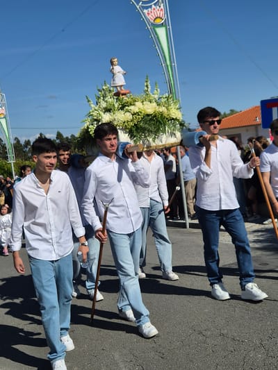 People wearing similar attire carry a religious float along a paved road. The float is decorated with flowers and a statue. The event appears to be taking place in Barcelos, Portugal, evidenced by the street sign in the background. The sky is blue.