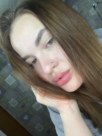 A close-up, angled, portrait-style photograph shows a fair-skinned individual with light brown, straight hair. The person's face is the main focus, featuring full, pink lips, a pierced nose, and light green eyes. The person's right hand, adorned with a ring, is positioned near their face. The individual is wearing a gray shirt. The background is a patterned surface. There is no visible location in the city of Port-Katon, Russia.