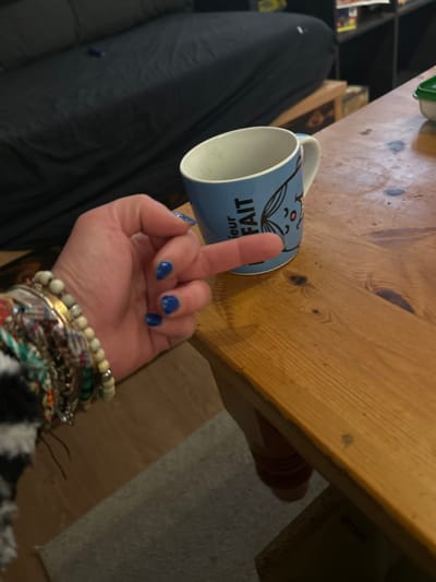 A blue mug with French text and a cartoon fox sits on a wooden table. A hand with blue nail polish extends its middle finger directly above the mug, obscuring part of the design. The wrist is adorned with numerous bracelets of various materials. Part of a black and white patterned sleeve is visible. The background includes a dark-colored cushioned chair and a portion of a floor.