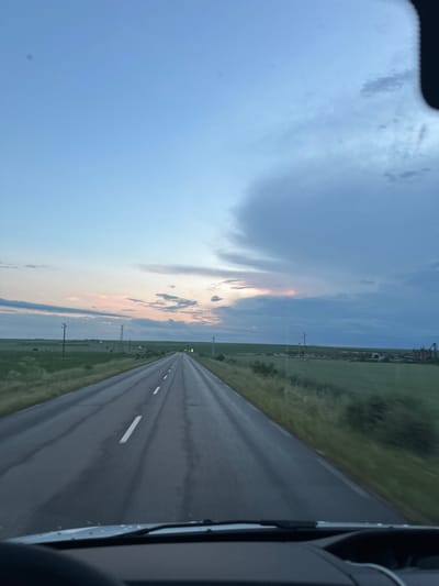 The image presents a view from a vehicle traveling along a straight, asphalt road, potentially near Zimnitsa, Bulgaria, though specific city elements are not visible. The road is bordered by grassy fields on either side, receding into the distance. Utility poles are spaced along the roadside. The sky is partly cloudy, with a mix of blue and areas of light orange and pink near the horizon, possibly indicating sunrise or sunset. The road's surface appears slightly wet, reflecting the ambient light. The passenger side of the vehicle is visible on the right.