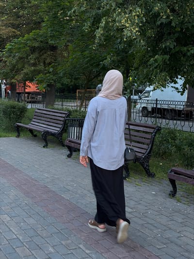 A person wearing a light beige hijab, a long-sleeved light blue shirt, and a black maxi skirt walks on a cobblestone path in what appears to be a park in Moskovskiy, Russia. The person is carrying a small black purse with a strap over their shoulder. In the background, there are dark wooden benches with ornate black metal legs, a trash receptacle, green bushes, and trees. Beyond the park area, there is a fence, a road with traffic, and a white truck. The lighting suggests it is daytime, likely late afternoon or early evening.