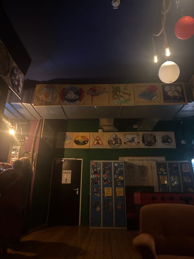 The image depicts the interior of a dimly lit establishment, possibly a bar or pub, in Leipzig, Germany. The scene is characterized by the presence of various artworks and decorations displayed on the walls.
On the upper wall, a row of square panels features illustrations, some with German text. One panel shows a bat-like creature, another a frog, and others depict animals such as a polar bear and sheep. Further along, a white spherical lamp and a red spherical decoration hang from the ceiling, suspended by a thick rope.
Below this, another row of square panels showcases different illustrations. These include a deer with a warning sign, a raccoon, a bird, a cat, and a dog. Beneath these artworks, a black door with a "TOILETS" sign is visible, indicating a restroom facility.
To the right of the door, a set of blue lockers is adorned with numerous stickers and badges. Next to the lockers, a red foosball table is partially visible. In the foreground, a portion of a plush, orange armchair is present, casting a shadow on the wooden floor.
The overall ambiance is subdued, with artificial lighting contributing to the dark and moody atmosphere. The details suggest a casual and potentially artistic environment.
