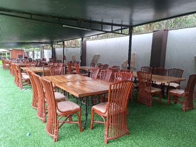 The image shows an outdoor dining area at a restaurant in Karagita, Kenya, located at coordinates -0.754 latitude and 36.437 longitude. The area is covered by a large awning supported by metal beams. The tables and chairs are made of wood and are arranged in rows under the awning. The chairs are brown wicker and the tables are dark wood with a light wood finish. The floor is covered in artificial green grass. There are no people present in the image. The restaurant is located in a suburb of Nairobi. It is unclear what time of day it is or if the restaurant is open.