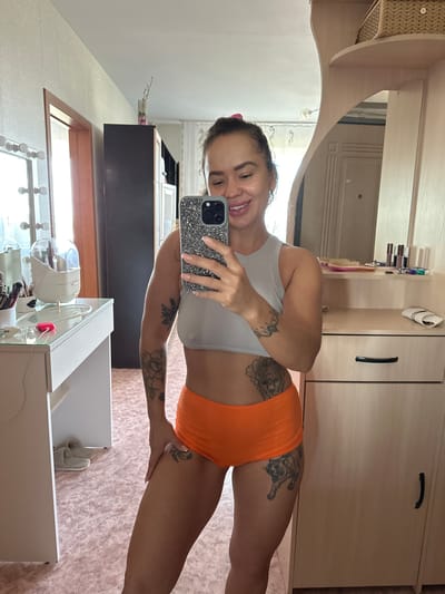 A woman is taking a mirror selfie in what appears to be a bedroom or dressing area. She is wearing an orange bottom and a grey top, and has tattoos on her legs and arm. She is smiling and holding up a phone with a sparkly case. In the background, there is a mirror reflecting a dresser with various items on it, including makeup and a wicker basket. A closet with a dark wooden door is also visible. The room has light pink patterned wallpaper. The lighting suggests it is daytime.