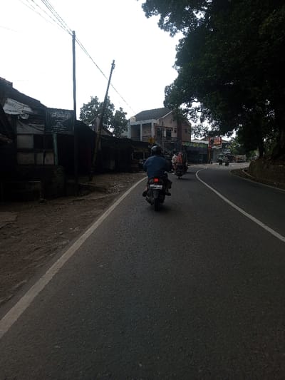 The image displays an asphalt road scene in the city of Chhenangri_Rishor, Bhutan, captured under an overcast sky. The road, appearing to accommodate at least two lanes, curves gradually to the right and is delineated by a solid white line on its left shoulder and a dashed white line further to the right, indicating traffic separation.
In the foreground, the back of an individual wearing a dark blue shirt and a black helmet is visible, operating a dark-colored scooter with license plate DK 7175 YH. This scooter is moving away from the viewer. Ahead of this scooter, a second individual on another scooter, carrying a large, bundled object behind the rider, also proceeds in the same direction. Further down the road, several additional vehicles, including other motorcycles and potentially a small truck, are observed following the road's curve.
To the left of the roadway, a dirt shoulder transitions into a series of structures. The nearest building appears dark and in disrepair, constructed with corrugated metal panels and partially covered by fabric or a tarp. A dark sign with white text is affixed to a pole in front of this structure. Beyond it, various other buildings, some appearing residential with multiple stories and light-colored facades, line the road. Interspersed among these buildings are trees and sparse vegetation. The right side of the road is bordered by an elevated, heavily vegetated embankment covered with large, dark-leafed trees that cast shadows across the road surface.
