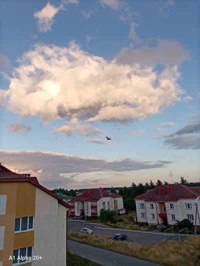 The image captures a scene in a residential area, likely in the Улукаўскі сельскі Савет, Belarus, during what appears to be late afternoon or early evening. A large, fluffy white cloud dominates the upper portion of the sky, with patches of blue visible around it. A dark bird is in flight in the middle ground of the sky. Below the sky, several multi-story apartment buildings with red roofs are visible. The buildings are constructed with light-colored brick and feature numerous windows. In front of the buildings, a paved road or driveway curves through a grassy area. Two cars are parked on the road. A tall, thin antenna is mounted on one of the buildings. The foreground features a portion of a yellow and cream-colored building with visible windows, and the text "A1 Alpha 20+" is superimposed in the lower left corner. The overall impression is of a typical urban or suburban residential setting under a partly cloudy sky.