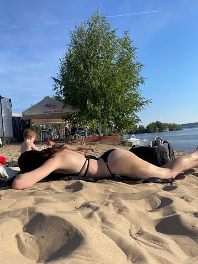 The image shows a woman in a black bikini lying on a sandy beach in Матюшино, Russia. She is lying on her stomach with her arms extended above her head. To her left is the beach, and to her right are a lake, trees, and a building that appears to be a café or restaurant. Other people are visible in the background. The sky is blue with a few clouds.