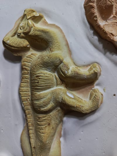A close-up shot displays a molded, three-dimensional representation of an anthropomorphic creature with reptilian and muscular human-like features, possibly made of clay or a similar material. The figure is tan and appears to be embedded in a white, plaster-like surface. The location is unknown; the image doesn't provide sufficient information to locate it in Guayaquil, Ecuador or anywhere else.