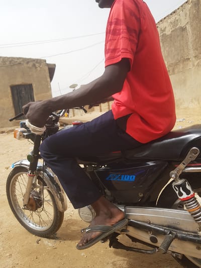 An individual wearing a red shirt and dark blue pants is seated on a Suzuki motorcycle, model AX100. The person is wearing a grey flip-flop on their right foot. The left hand is gripping the handlebar, which is wrapped with a white bandage. The setting appears to be Dutsinma B, Nigeria, suggested by the surrounding architecture. A building with a window is visible in the background. The ground appears to be dirt.