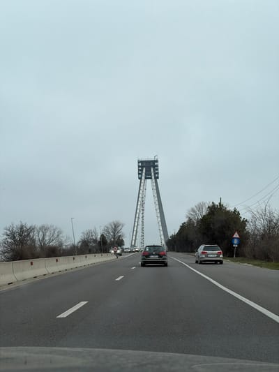 The image presents a forensic view of a multi-lane asphalt highway leading towards a prominent bridge tower structure in Agigea, Romania, under an overcast grey sky. The perspective is from within a moving vehicle, indicated by the dashboard or lower windshield edge at the bottom of the frame.

In the immediate midground, a dark grey Volkswagen Golf hatchback (Romanian license plate CT 13 MZA) occupies the left lane, followed by a silver BMW 5 Series Touring wagon (Romanian license plate CT 07 BWN) in the right lane. Multiple additional vehicles are visible in the distance, proceeding towards the bridge.

The central subject is a tall, modern, inverted-V-shaped bridge pylon, constructed from grey metallic components, featuring a rectangular enclosure at its apex and visible access ladders or structural supports running along its inner sides.

To the left of the highway, a concrete barrier delineates the road from an embankment populated by bare deciduous trees and streetlights. To the right, a dirt verge borders the road, displaying a mix of bare trees and dense evergreen shrubs. Overhead utility lines are visible against the sky. A blue and white circular road sign, signifying "no stopping" or "no parking" (with a red diagonal line through a white 'P' symbol), is positioned on the right verge, beneath which is a smaller blue circle with a white '0'. The highway surface features dashed white lane markings and a solid white line defining the right shoulder. No identifiable individuals are visible within the scene.