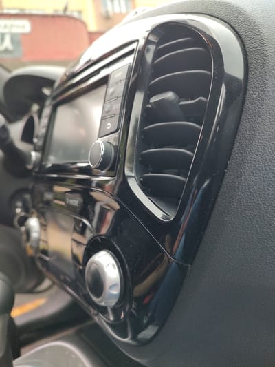 The image shows a close-up view of a car's center console, specifically the infotainment system and an air vent. The console is made of black glossy plastic and textured black material. On the left side of the image, a portion of the screen of the infotainment system is visible, along with several buttons. One button labeled "MAP" is clearly legible. Below the screen, there are knobs and buttons, one of which is a silver-colored rotary dial with a textured grip. To the right of the infotainment system is a prominent air vent with adjustable louvers, framed by a glossy black bezel. The background is out of focus, showing hints of buildings and possibly a street scene, suggesting the car is parked outdoors. The lighting indicates it is daytime, with natural light illuminating the scene. There are no people visible in the image. The scene appears to be a still shot of the interior of a car, likely a Nissan Juke based on the design elements. The location in the city of Tervel, Bulgaria, is not visually identifiable from the provided image alone.